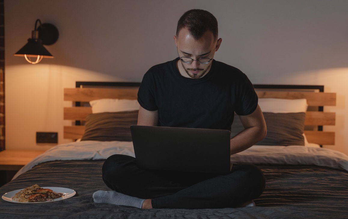 A young man in glasses sitting cross-legged on a bed at night, working on a laptop